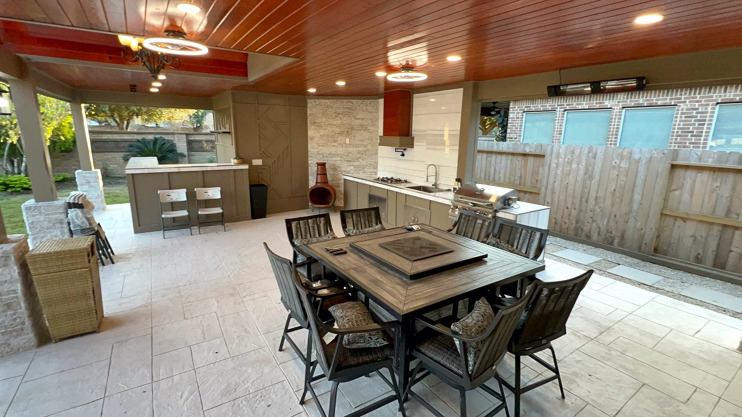 Interior of covered outdoor kitchen showing stained wood ceiling, gas cooking station, dining table, and large-format tile fl