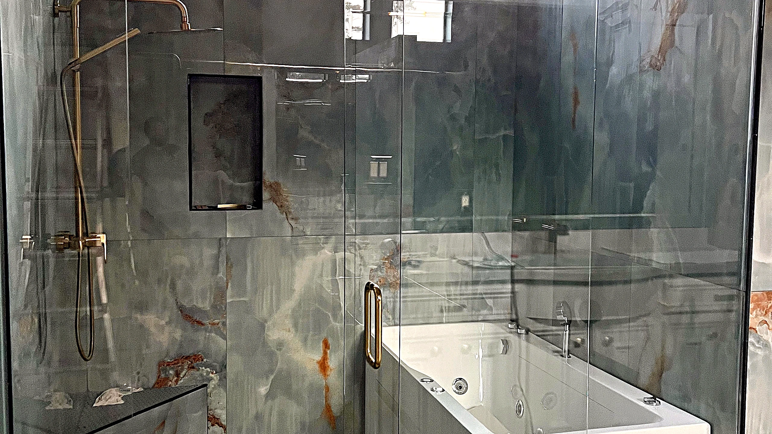 Natural stone slab shower interior with brushed-gold rain shower system, recessed niche, and white soaking tub behind framele