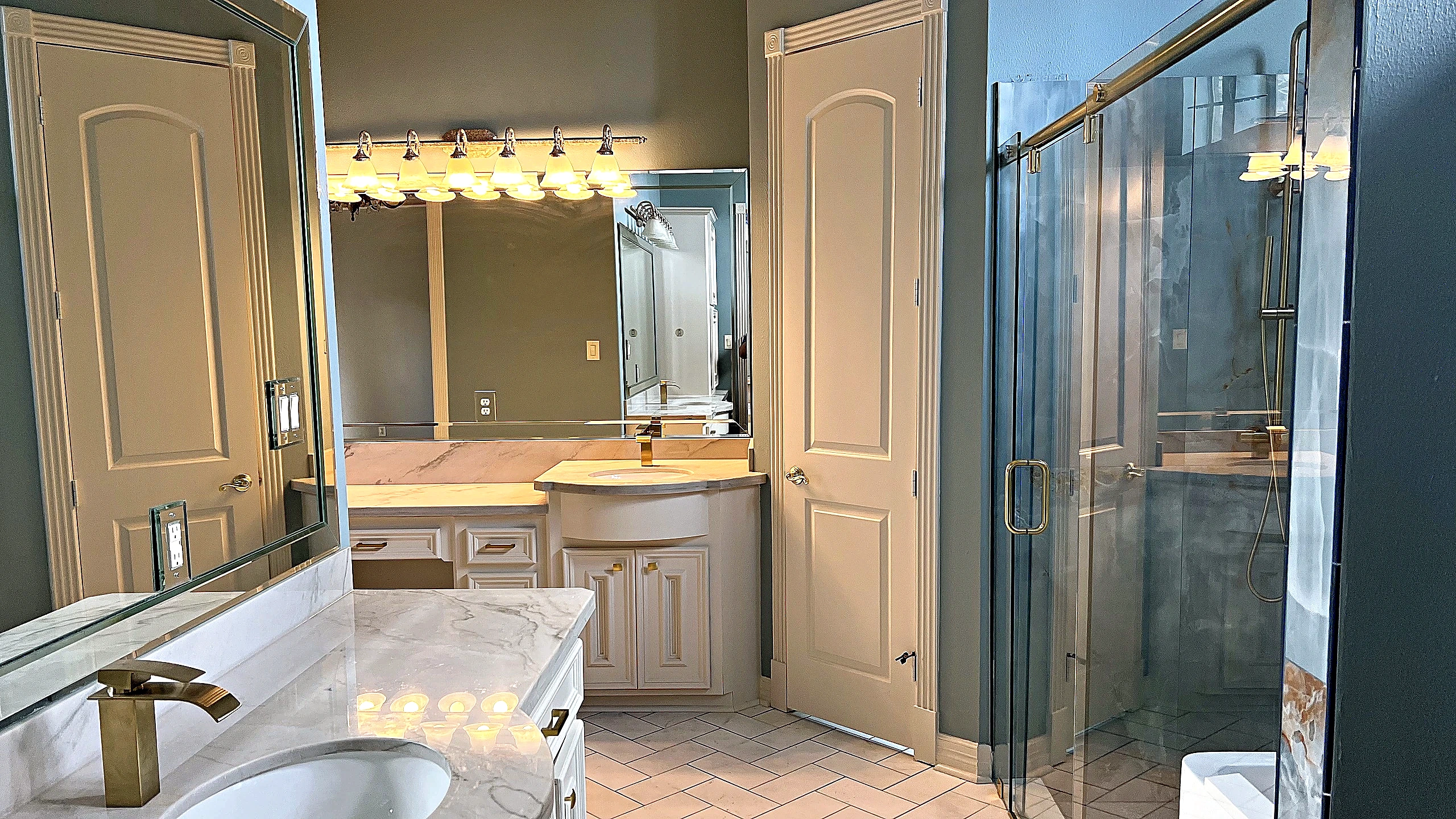 Renovated master bathroom vanity area with white cabinetry, veined stone countertop, gold faucets, and view to glass-enclosed