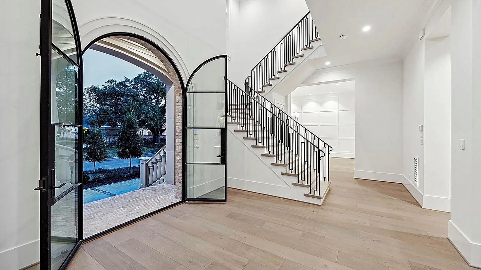 Completed River Oaks entry with arched steel-and-glass pivot door open, wide-plank wood floors, and curved iron-railed stairc