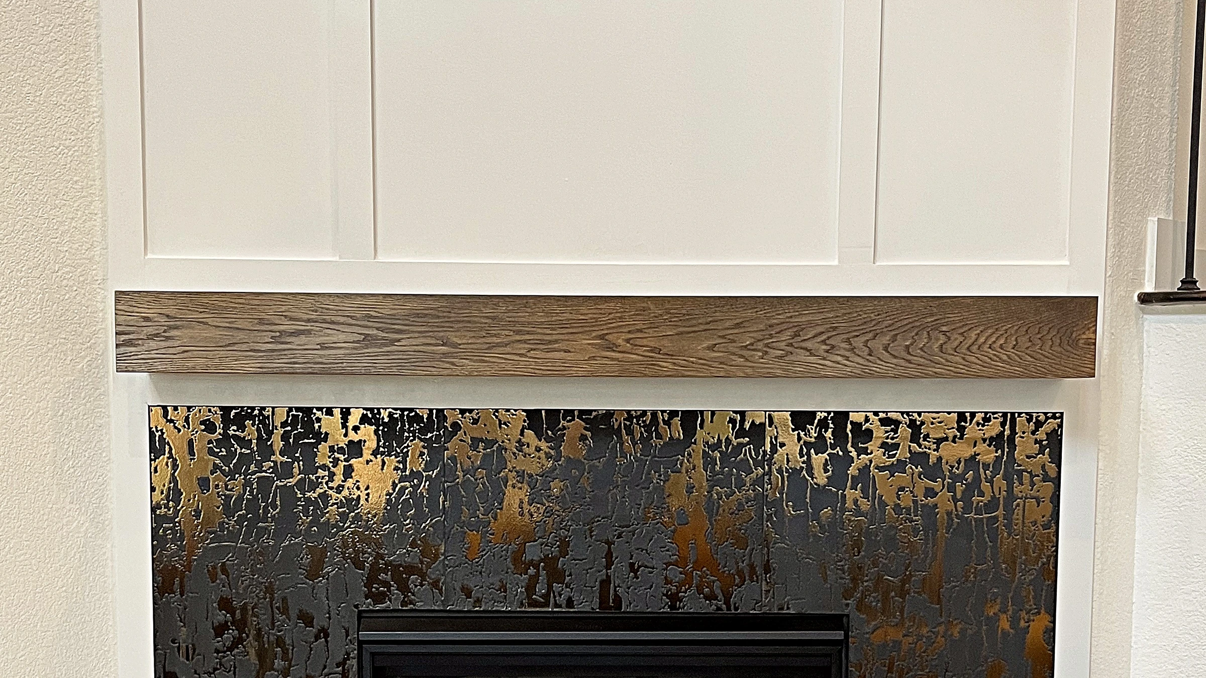 Close detail of floating dark-stained wood mantel shelf above textured charcoal-and-gold tile fireplace surround